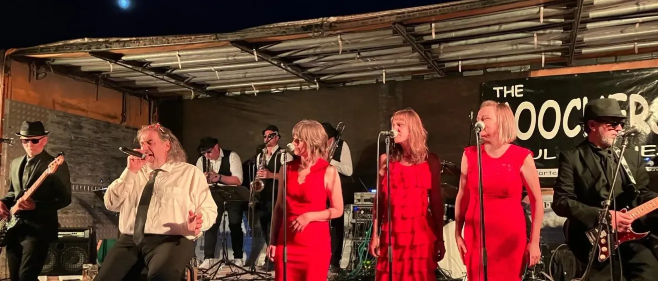 Eine Band tritt bei Nacht auf der Bühne auf, mit zwei Frauen in roten Kleidern, die singen, und anderen, die Instrumente spielen.
