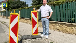 Ein Mann in einem weißen Hemd und Jeans steht neben zwei roten und weißen Verkehrszeichen an einer Straße mit einem Steinbord und einem grünen Zaun.