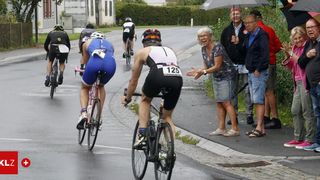Eine Gruppe von Radfahrern in einem Rennen mit Zuschauern, die jubeln. Eine ältere Frau spricht mit einem Radfahrer mit der Nummer 125.