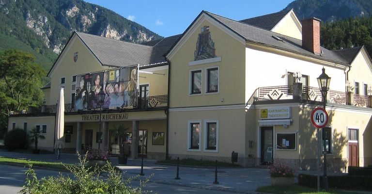 Das Reichenau Theater zeigt ein Wandbild auf seiner Fassade, mit Pflanzen und Straßenpollern davor. Das Theaterladen-Schild ist am Gebäude zu sehen.