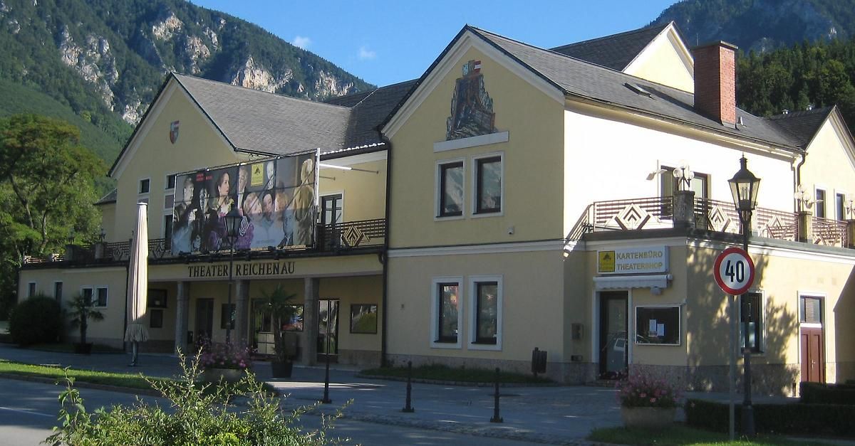 Das Reichenau Theater zeigt ein Wandbild auf seiner Fassade, mit Pflanzen und Straßenpollern davor. Das Theaterladen-Schild ist am Gebäude zu sehen.