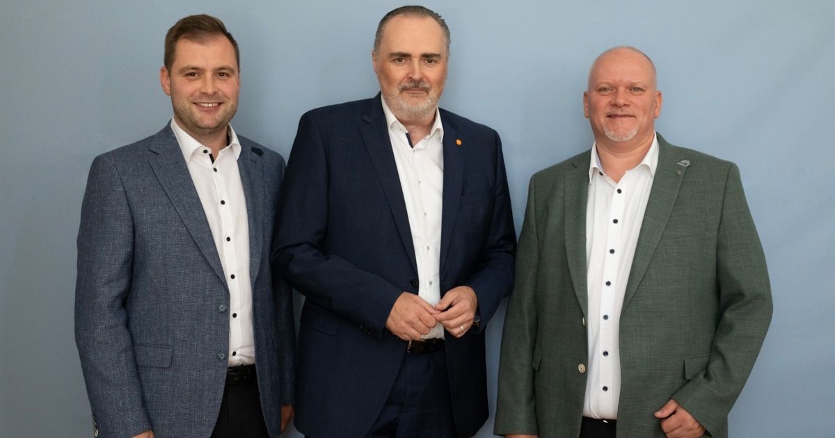 Drei Männer in Anzügen und weißen Hemden stehen zusammen vor einer blauen Wand und lächeln für die Kamera.