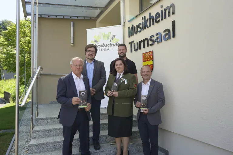 Fünf Personen stehen auf Stufen vor einem Gebäude mit der Aufschrift 'Musikheim Turnsaal'. Sie halten Bücher in den Händen und lächeln.