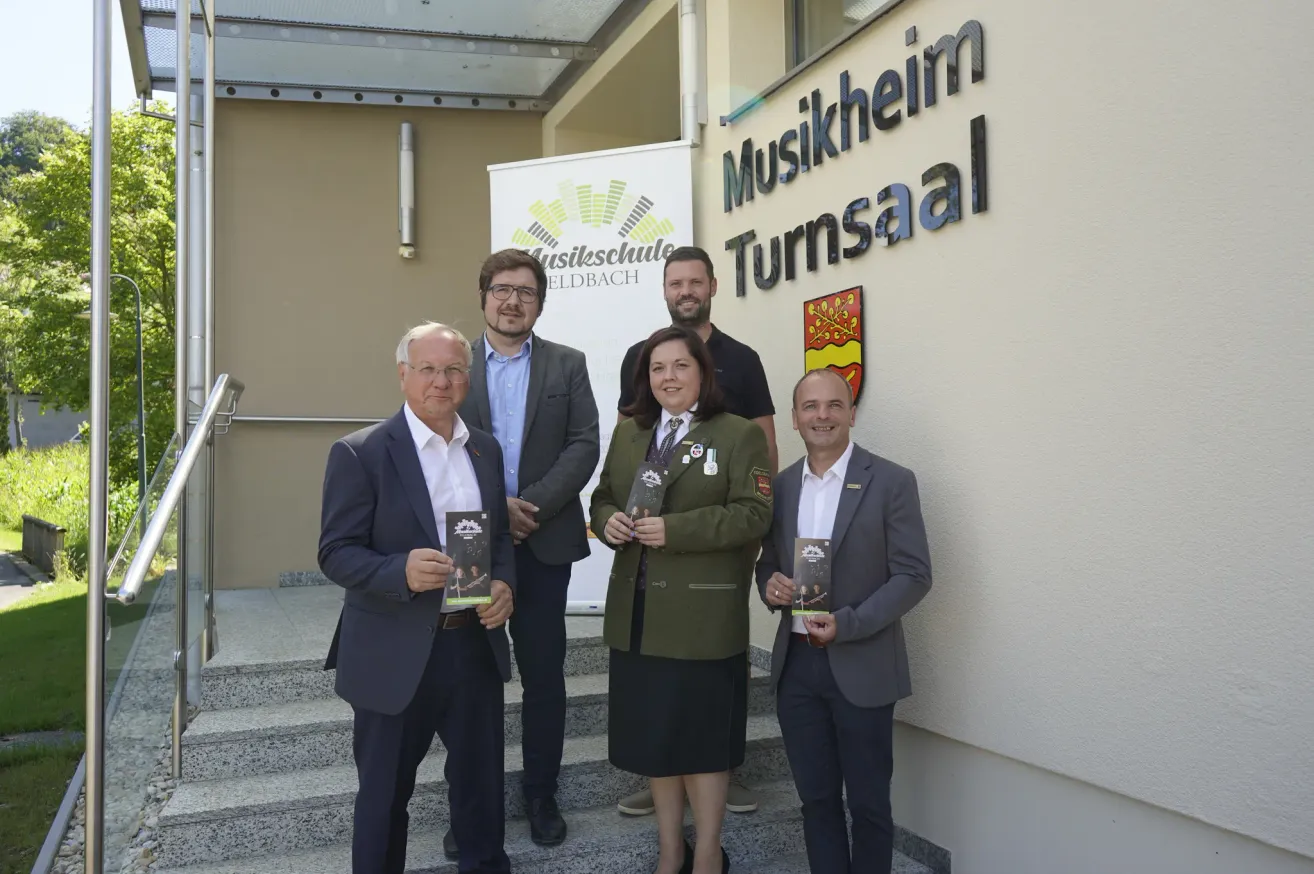 Fünf Personen stehen auf Stufen vor einem Gebäude mit der Aufschrift 'Musikheim Turnsaal'. Sie halten Bücher in den Händen und lächeln.