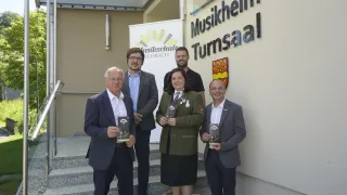 Fünf Personen stehen auf Stufen vor einem Gebäude mit der Aufschrift 'Musikheim Turnsaal'. Sie halten Bücher in den Händen und lächeln.