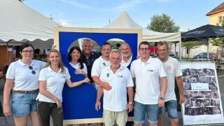 Eine Gruppe von Menschen, die weiße Hemden mit Logos tragen, posiert hinter einem großen blauen herzförmigen Ausschnitt und lächelt für die Kamera. Sie stehen unter einem blauen Himmel.