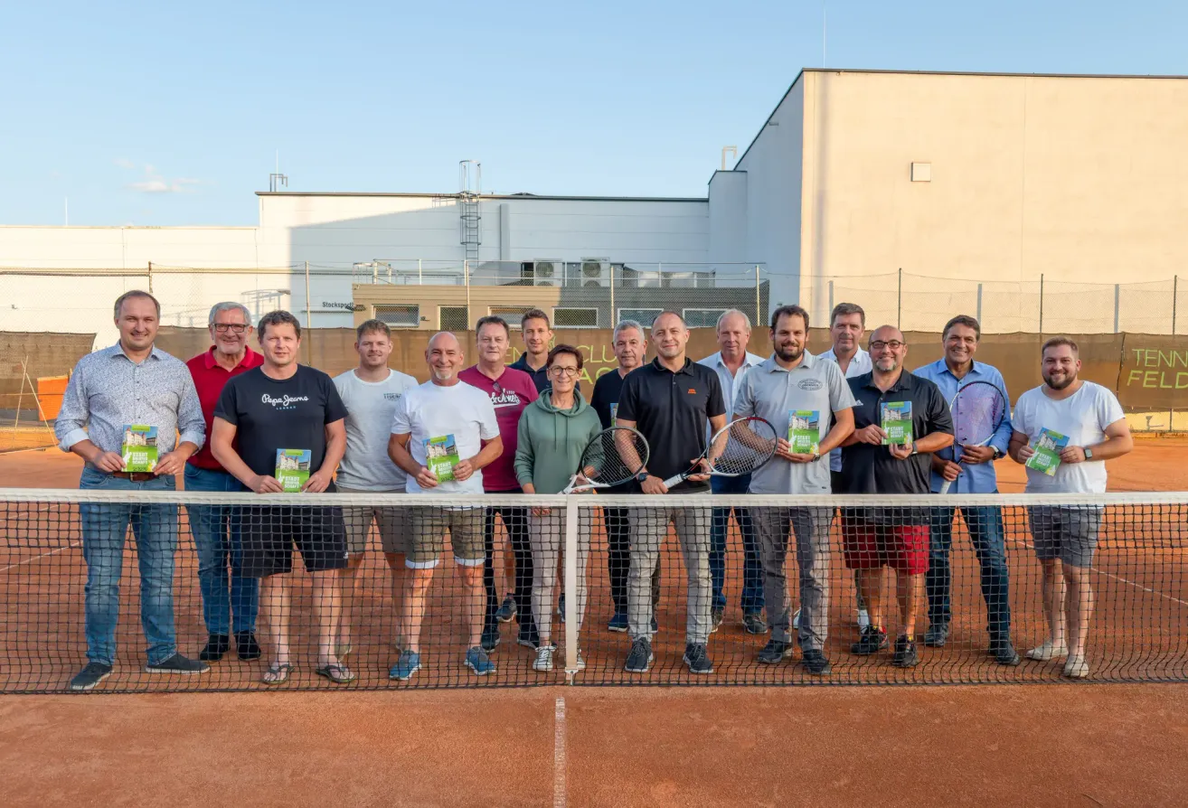 Eine Gruppe von Menschen in Sportkleidung steht hinter einem Tennisnetz mit Tennisschlägern und Büchern in der Hand, mit Gebäuden im Hintergrund.