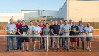 Eine Gruppe von Menschen in Sportkleidung steht hinter einem Tennisnetz mit Tennisschlägern und Büchern in der Hand, mit Gebäuden im Hintergrund.