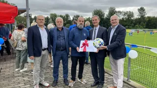 Fünf Männer stehen nebeneinander auf einem Ziegelweg, einer hält einen Fußball. Sie scheinen eine Auszeichnung zu überreichen. Der Hintergrund zeigt ein Fußballfeld mit einem Netz und Luftballons.