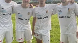 Vier Männer in weißen Uniformen posieren für ein Foto auf einem Rasenfeld. Sie lächeln für die Kamera. Die Uniformen haben roten und weißen Text und Zahlen. Der Hintergrund umfasst Berge und ein Schild.