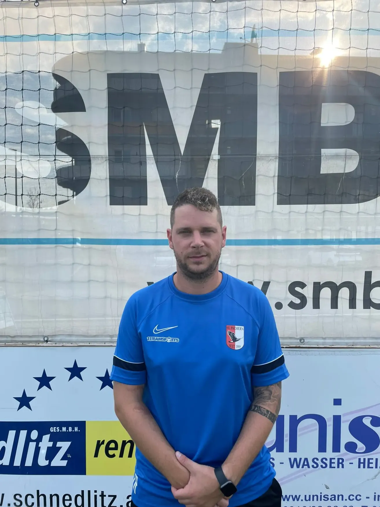 Ein Mann in einem blauen Shirt mit Logo steht vor einem Banner mit dem Text SMB. Das Banner zeigt auch ein Netz und Sterne.