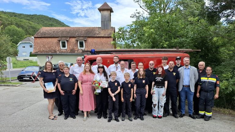 Eine Gruppe von Feuerwehrleuten posiert für ein Foto vor einem Feuerwehrauto und einem Gebäude mit einem Turm. Einige Personen tragen Uniformen, während andere Freizeitkleidung tragen.
