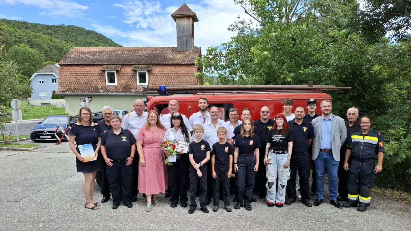Eine Gruppe von Feuerwehrleuten posiert für ein Foto vor einem Feuerwehrauto und einem Gebäude mit einem Turm. Einige Personen tragen Uniformen, während andere Freizeitkleidung tragen.