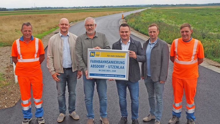 Vier Männer stehen auf einer neuen Straße mit einem Schild, das 'Für mehr Sicherheit - Bahnübergang Absorf - Utzenlaa' steht. Sie sind in Geschäftskleidung gekleidet und lächeln für die Kamera.