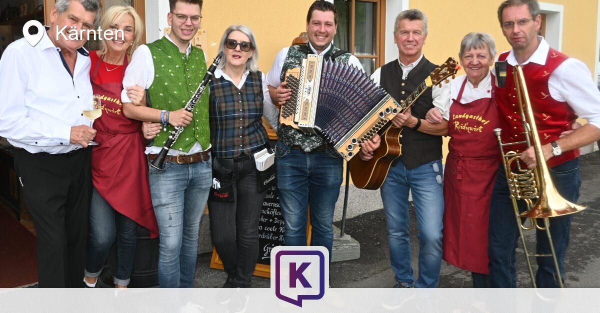 Eine Gruppe von Musikern in traditioneller Kleidung posiert für ein Foto. Einer hält ein Akkordeon, ein anderer eine Klarinette und ein Dritter eine Gitarre. Hinter ihnen steht eine Tafel mit deutscher Beschriftung.