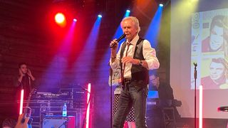Ein Mann steht auf der Bühne und singt in ein Mikrofon, trägt eine Weste und Jeans. Hinter ihm steht eine Frau in der Nähe einer Gitarre, und ein Mann spielt Klavier. Scheinwerfer und Lautsprecher sind auf der Bühne aufgestellt.