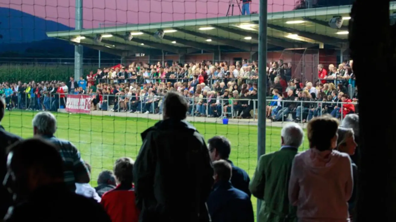 Eine Menschenmenge versammelt sich in einem Stadion für eine Veranstaltung, einige stehen, andere sitzen. Die Menschen tragen verschiedene Farben und Kleidung. Das Spielfeld ist von einem Zaun umgeben, und ein Tornetz ist sichtbar.