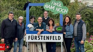 Eine Gruppe von Menschen posiert für ein Foto in einem Garten mit einem Schild, auf dem 'Fürstenfeld' steht. Eine Frau hält einen Korb und ein Mann eine Gießkanne. Über ihnen befindet sich eine Sprechblase mit dem Text 'Ich will ham noch'.