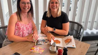 Zwei lächelnde Frauen sitzen an einem Tisch mit Zeitschriften, einem Handy und Gewürzen auf einer Terrasse. Sie sind in der Nähe einer Glaswand mit Blick auf Autos und Grünflächen.