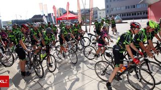 Eine Gruppe von Radfahrern in passenden grünen und schwarzen Uniformen fährt auf einer gepflasterten Fläche mit Gebäuden im Hintergrund.