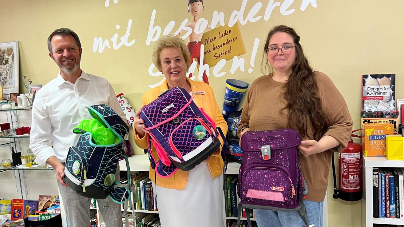 Drei Personen stehen in einer Bibliothek. Zwei von ihnen halten Rucksäcke. Die Frau in der Mitte lächelt und hält einen Rucksack. Hinter ihr steht ein Regal mit Büchern. Die Frau rechts trägt eine Brille und hält einen Rucksack.