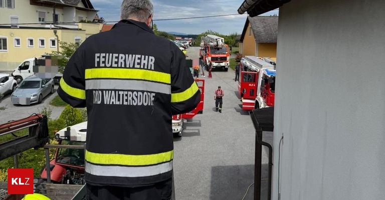 Ein Feuerwehrmann von Rad Waltersdorf steht auf einem Parkplatz mit Feuerwehrwagen. Menschen sind versammelt, möglicherweise im Einsatz bei einem Notfall.