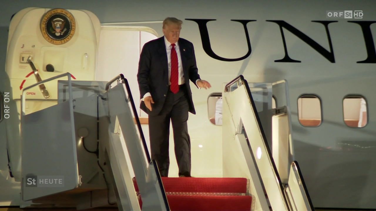 Donald Trump steigt von einem Flugzeug aus und trägt einen schwarzen Anzug, eine rote Krawatte und Lederschuhe. Er hält eine Mappe in der linken Hand.