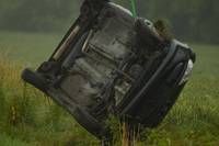 Ein umgekipptes Auto liegt auf der Seite in einem Grasfeld. Der Karosserie des Autos ist schwarz, und die Reifen sind platt. Ein grünes Seil ist am Auto befestigt, was darauf hindeutet, dass es aus dem Gras gezogen wurde.