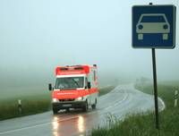 Ein Krankenwagen fährt auf einer nebligen Straße, auf der rechten Seite befindet sich ein Schild mit einem Auto.