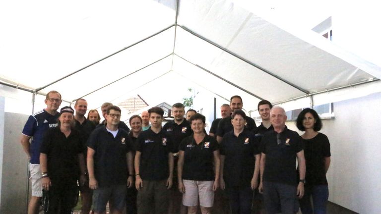 Eine Gruppe von Menschen in schwarzen Uniformen mit weißen Logos steht unter einem weißen Zelt. Sie lächeln für ein Foto.