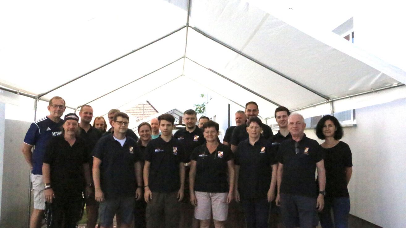 Eine Gruppe von Menschen in schwarzen Uniformen mit weißen Logos steht unter einem weißen Zelt. Sie lächeln für ein Foto.