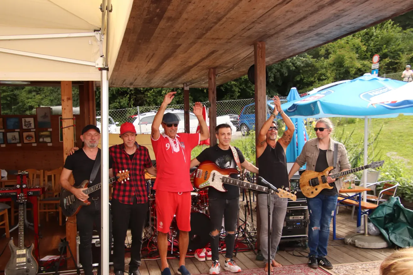 Eine Gruppe von Musikern tritt auf einer Bühne auf. Der Mann im roten Shirt hebt seine Hände. Es gibt Gitarren, ein Schlagzeug und ein Mikrofon auf der Bühne.