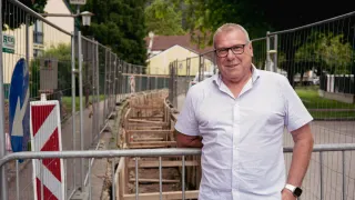 Ein Mann in einem weißen Hemd und mit Brille steht neben einer Baustelle an einer Straße mit Metallzäunen. Bäume und Häuser sind im Hintergrund.