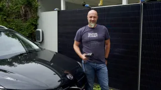 Ein Mann in einem T-Shirt mit der Aufschrift 'Because there is no planet B' lädt ein Elektroauto mit einem Ladekabel auf. Er steht vor einem Solarpanel.