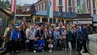 Eine Gruppe älterer Menschen posiert für ein Foto auf einer Stadtstraße mit Geschäften und Gebäuden im Hintergrund. Einige tragen Stöcke und ein junges Mädchen hält ein Kreuz.