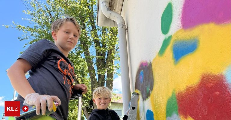 Zwei junge Jungen stehen auf Leitern und malen ein farbenfrohes Wandbild an der Seite eines Gebäudes. Einer hält einen Pinsel und Sprüher, während der andere einen Pinsel hält. Ein Baum und ein Haus sind hinter ihnen.