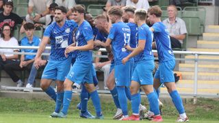 Fußballspieler in blauen Trikots feiern auf einem Feld mit Zuschauern im Hintergrund, einige tragen Hüte und Brillen.