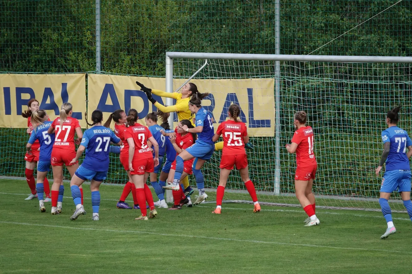 Weibliche Fußballerinnen in roten und blauen Trikots spielen auf einem Feld. Eine Spielerin in einem gelben Trikot springt, um ein Tor zu blockieren. Die Torfrau trägt ein blaues Trikot mit der Nummer 14.