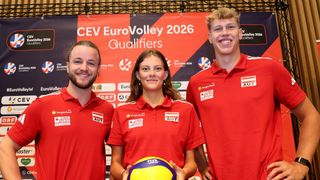 Drei Personen in roten Poloshirts stehen eng zusammen, lächeln und posieren für ein Foto vor einem Banner, das CEV EuroVolley 2026 Qualifikationen liest. Die Frau in der Mitte hält einen Volleyball.