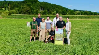 Eine Gruppe von Menschen steht in einem Feld mit einem Hund und einem Schild, das 'Bluhende und Summende Steiermark' sagt. Sie lächeln und scheinen für ein Foto zu posieren.
