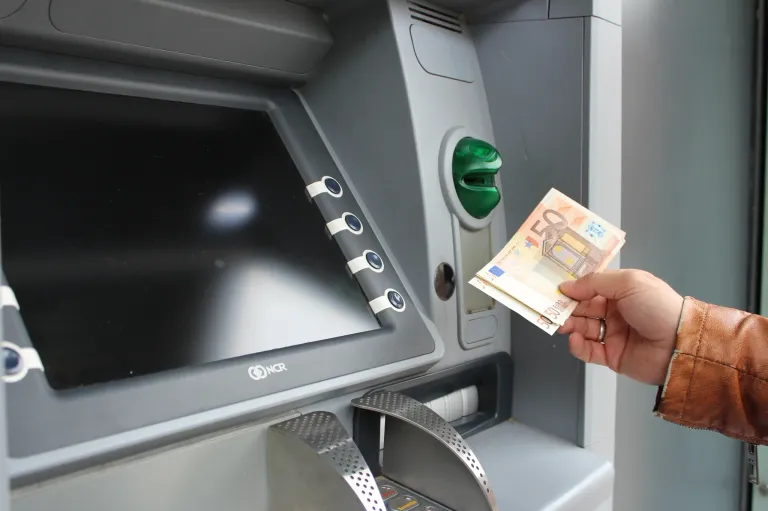 Eine Person steckt einen 50-Euro-Schein in einen Geldautomaten. Der Geldautomat ist grau mit einem großen Bildschirm und Tasten.