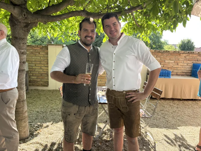 Zwei Männer stehen lächelnd unter einem Baum, einer in einem weißen Hemd und der andere in traditioneller Kleidung, mit einem Glas Bier in der Hand.