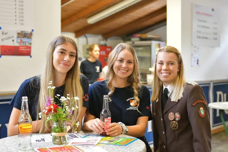 Drei lächelnde junge Frauen sitzen mit Blumen und einer Limonadenflasche an einem Tisch in einem Raum mit Holzdecke.
