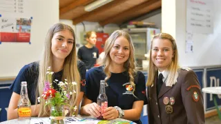 Drei lächelnde junge Frauen sitzen mit Blumen und einer Limonadenflasche an einem Tisch in einem Raum mit Holzdecke.