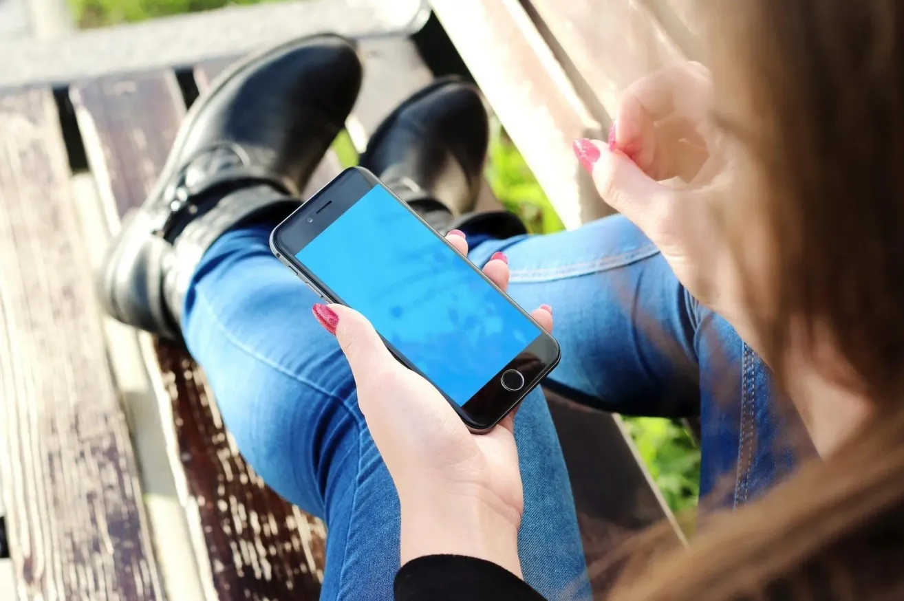 Eine Person in blauer Jeans und schwarzen Stiefeln sitzt auf einer Bank und hält ein schwarzes Handy mit blauem Bildschirm.