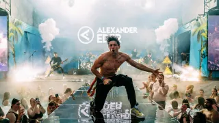 Ein oberkörperfreier Mann tritt auf einer Bühne mit Rauch- und Nebeleffekten auf. Hinter ihm spielt eine Band auf einer erhöhten Plattform mit einem Hintergrund, der das Alexander Eder-Logo zeigt. Das Publikum ist durchnässt.