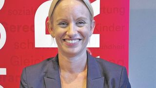 Eine Frau mit kurzen blonden Haaren, die einen dunkelblauen Blazer und ein gelbes Top trägt, lächelt fröhlich vor einem roten Hintergrund.