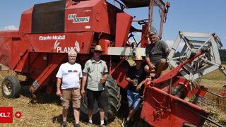 Vier Männer posieren für ein Foto neben einer roten Erntemaschine auf einem Feld. Ein Mann hält ein Glas mit Getränk.