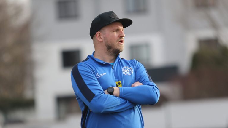 Ein Mann in einem blauen Jacke mit Nike-Logo, einer schwarzen Kappe und einer Uhr, steht im Freien mit verschränkten Armen.