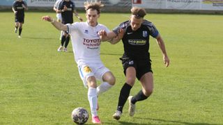 Zwei Fußballspieler konkurrieren auf einem Feld. Einer trägt ein weißes Toyota Buchereiten-Trikot, der andere ein schwarzes Eco Pule-Trikot. Beide sind in einen Zweikampf um den Ball verwickelt.